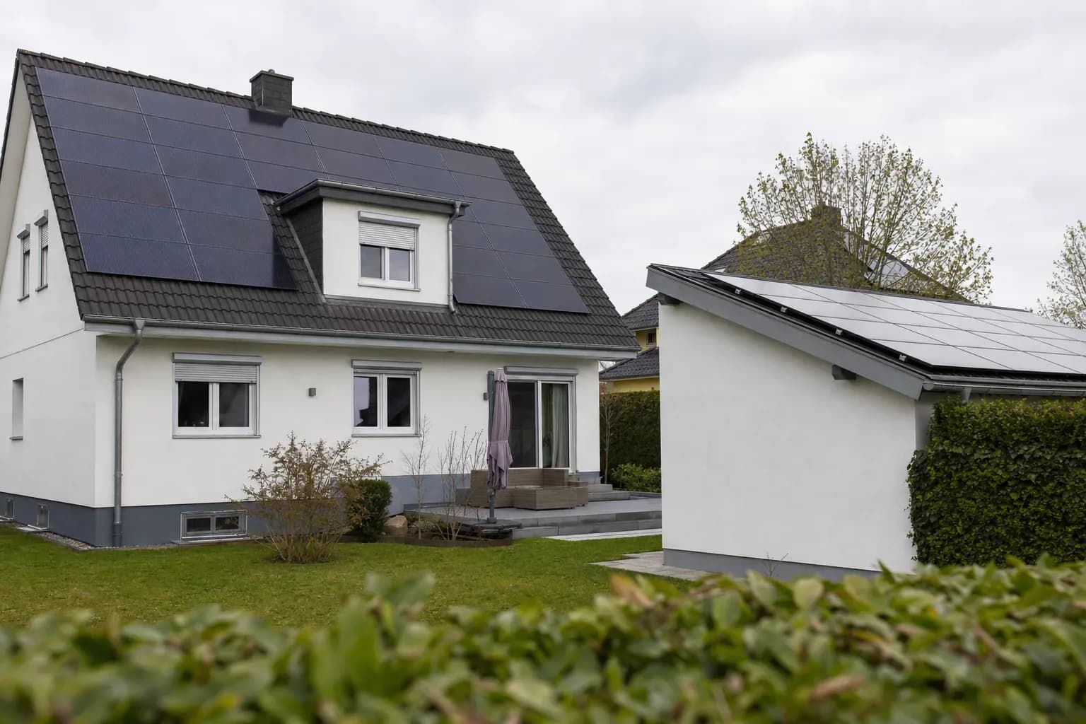 Hinterhofansicht mit dem Dach eines Wohnhauses und dem Dach einer freistehenden Garage, beide mit Photovoltaikmodulen, bei hellem bedecktem Tageslicht; Hecke und Nachbarhauskanten sind am Rand unscharf.