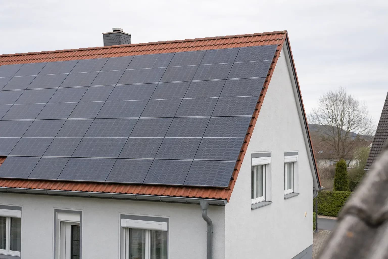 Schräger Blick von gegenüber auf das Ziegeldach eines Einfamilienhauses mit installierter Photovoltaikanlage; Nachbarhauskanten und Baumkronen sind unscharf, heller Himmel mit hoher Bewölkung.